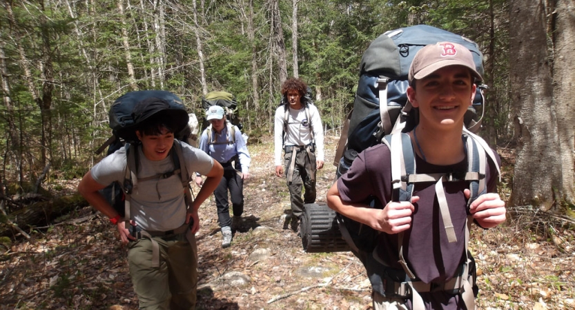 Four students wearing backpacks hike through a wooded area. 